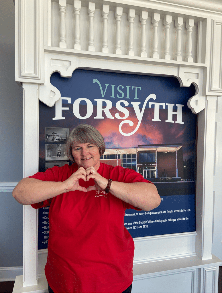 Abbie Bunn, a Tift College alumna, stands in front of a Visit Forsyth sign making a heart with her hands
