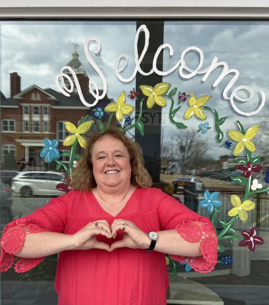 Life-long Forsyth Resident Dulcie Whitaker,  who is active in the 1823 Artisan Guild, stands in front of a window she painted at the Forsyth Welcome Center making a heart with her hands
