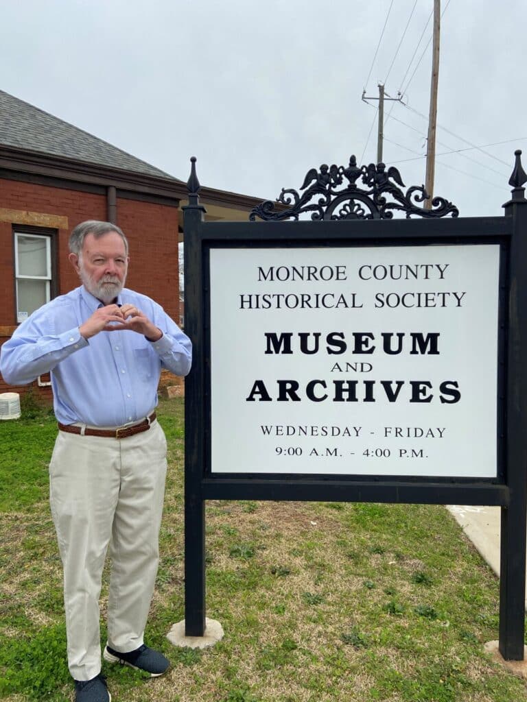 Local Historian Ralph Bass stands by the sign for the Monroe County Historical Society Museum making a heart with his hands