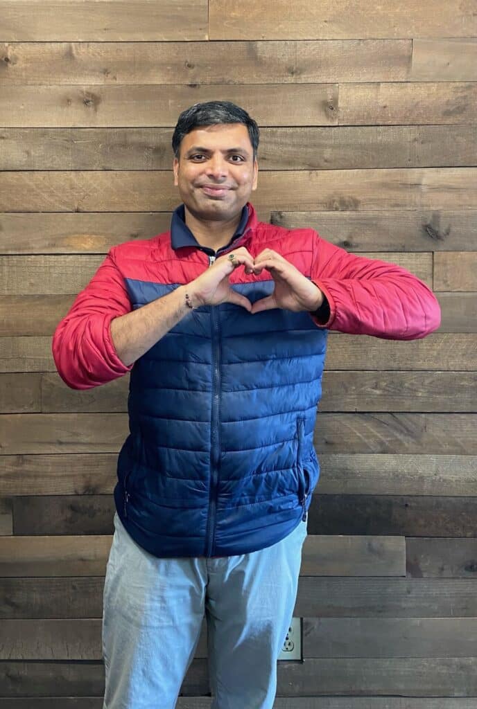 Sam Patel, owner of Forsyth Travelodge, stands in front of a wood panel lodge and makes a heart with his hands